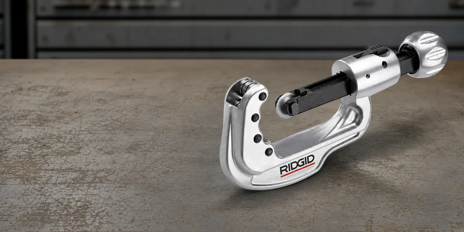 RIDGID stainless steel tubing cutter shown in a workshop setting on a worn metal surface, with close-up detail of the cutting wheel, adjustment screw and durable metal body.